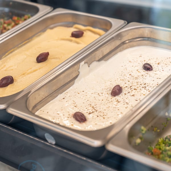 Creamy hummus and a seasoned white dip with olives in a restaurant display case.