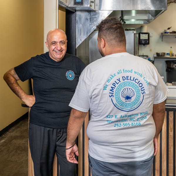 Smiling man in Simply Delicious shirt with staff member in their Greek restaurant kitchen.