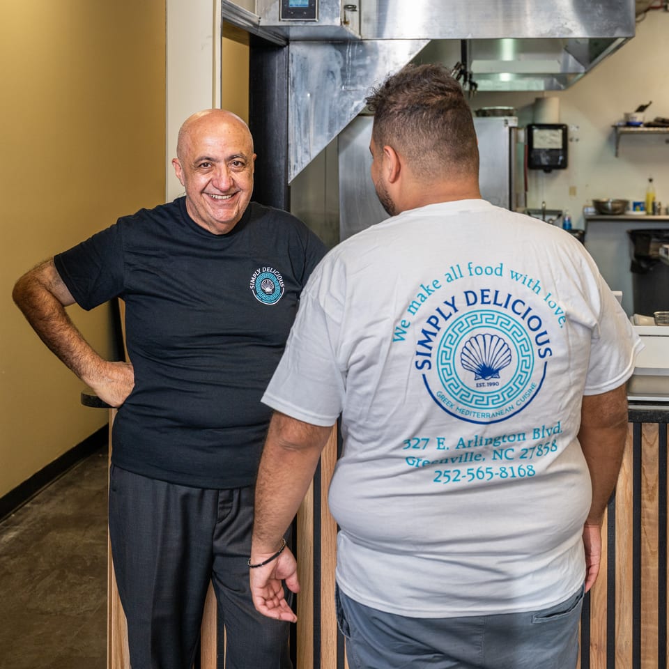 Smiling man in Simply Delicious shirt with staff member in their Greek restaurant kitchen.