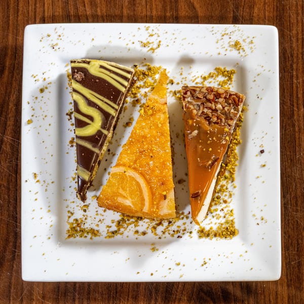 Three dessert slices: chocolate, orange, and caramel pecan, on a white plate with pistachio crumbs.