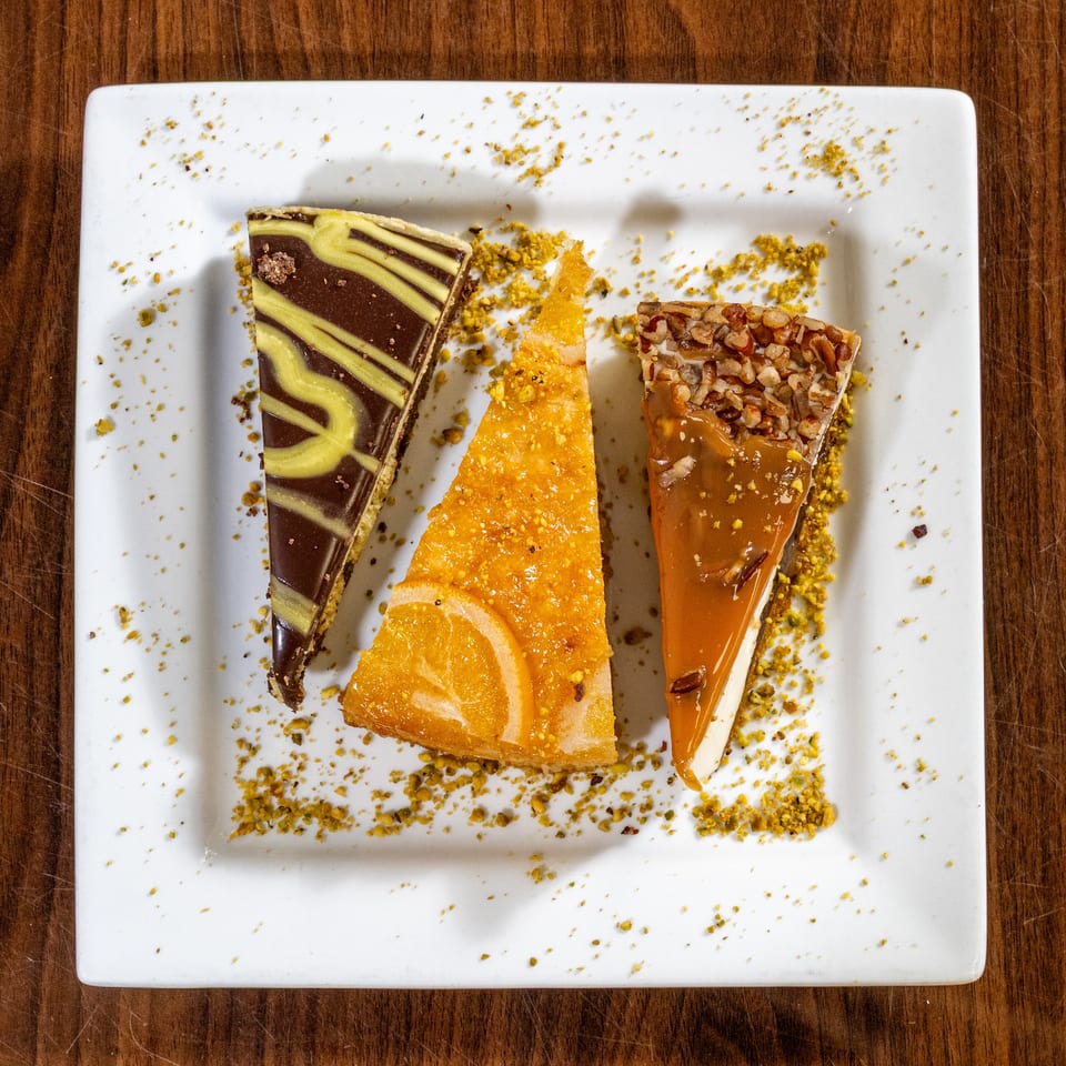 Three dessert slices: chocolate, orange, and caramel pecan, on a white plate with pistachio crumbs.