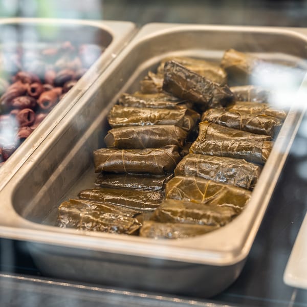 Fresh dolmades in a metal serving tray, with olives visible in the background.