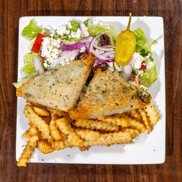 Golden spanakopita triangles, crinkle fries, and a Greek salad with feta and pepperoncini on a white plate.
