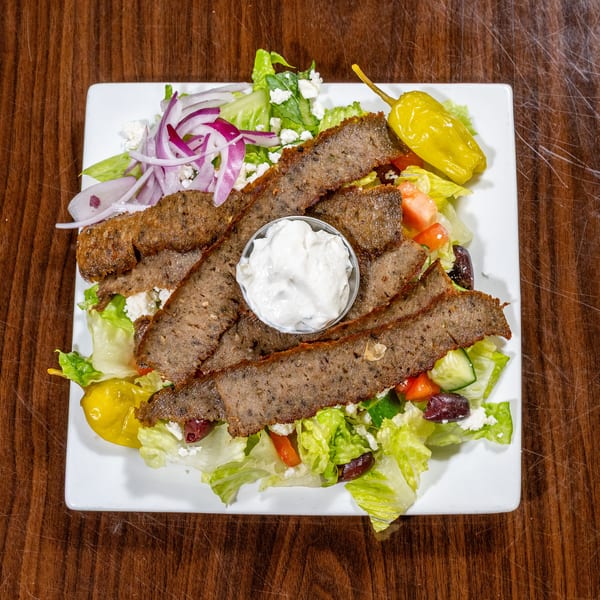 Gyro meat slices on a Greek salad with tzatziki, feta, and pepperoncini on a white plate.