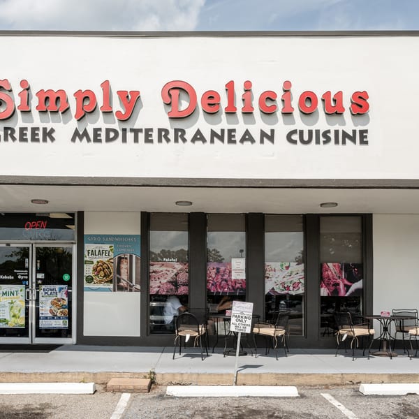 Simply Delicious Greek Mediterranean Cuisine storefront with outdoor tables and promotional window posters.