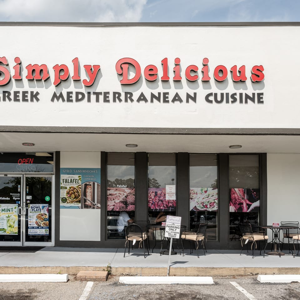 Simply Delicious Greek Mediterranean Cuisine storefront with outdoor tables and promotional window posters.