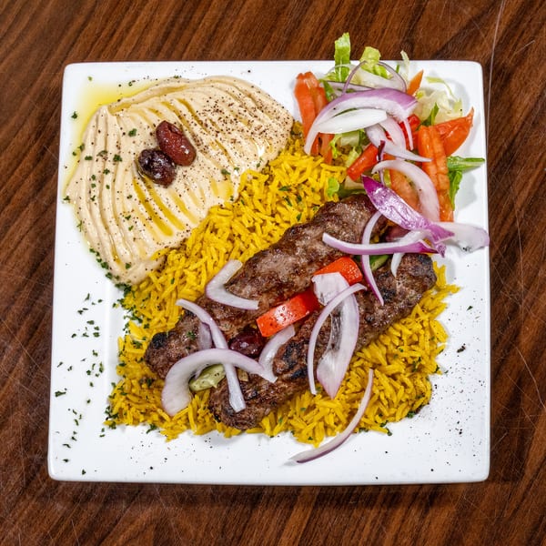 Grilled kofta kebab, yellow rice, hummus with olives, and a fresh side salad on a white plate.